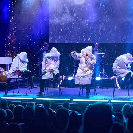 Four performers in long white coats with fur trim hoods sit on stools and sing.