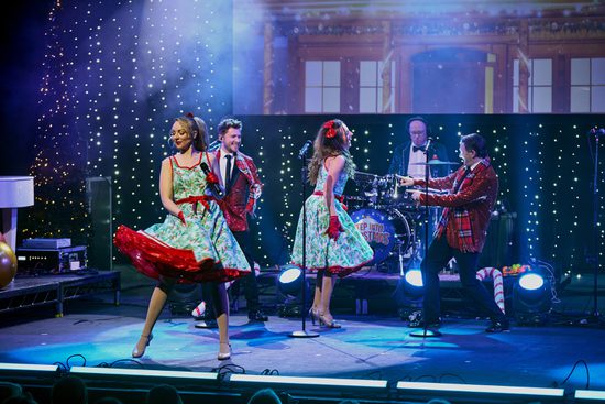 Two men in red and green suits dance with two women in dresses covered in holly.