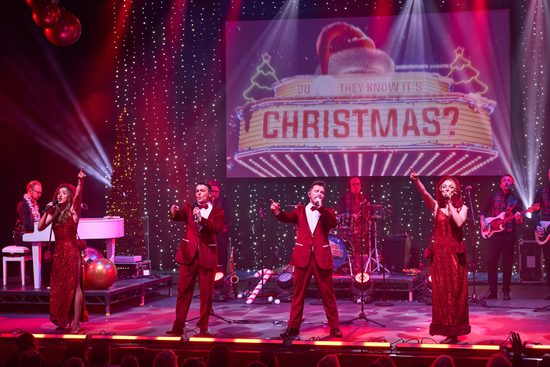 Two men in red suits and two women with red dresses with hoods singing on a stage with musicians behind them.