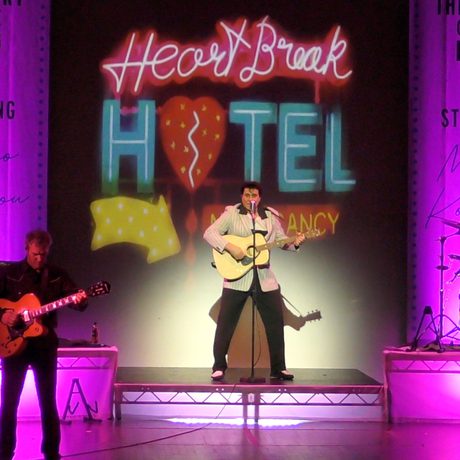 A man in a white pinstripe suit stands at the back of a purple-lit stage and plays a guitar. Behind him is a neon sign that says Heartbreak Hotel, and he is accompanied by various other musicians playing keyboard, guitar, drums, and double bass.