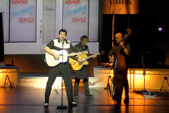 A brunette man in a black and white shirt plays a guitar on a stage and sings. He is accompanied by men playing guitar and double bass.