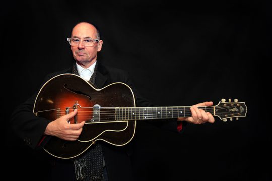 A bald bam wearing glasses and a black suit. He is holding a deep red guitar, standing against a black background.