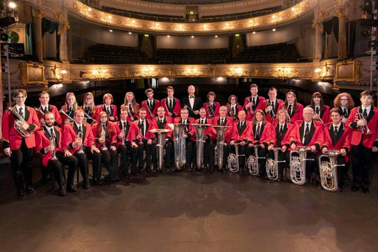 A brass band stood on a stage with an ornate auditorium in the background. They are holding their instruments and wearing red jackets and black ties.