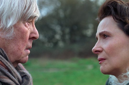 A man and a woman stand very close together, looking into each others eyes with concern. They are stood in a grassy field.