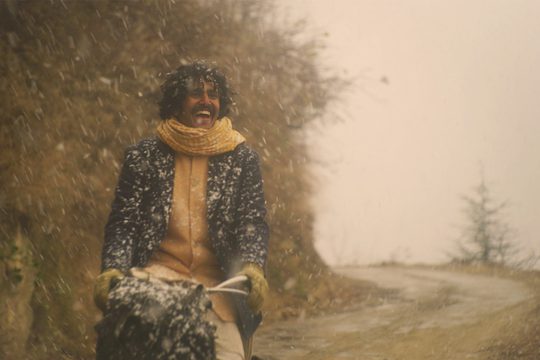 A man wearing a yellow shirt and navy blue jacket with a yellow scarf cycles in the snow. He is smiling widely.