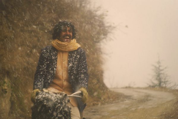 A man wearing a yellow shirt and navy blue jacket with a yellow scarf cycles in the snow. He is smiling widely.