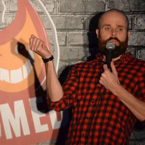 A man in a red and black checked shirt with a brown beard holds a microphone on stage.
