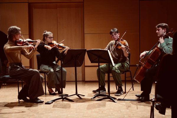 Four musicians playing violin, viola, and cello sat on chairs behind music stands. They are sat in a semicircle formation.