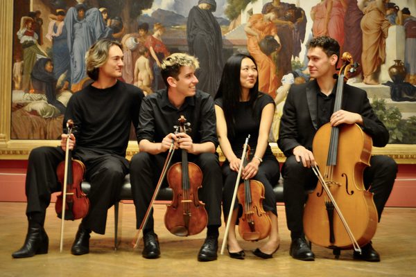 Four musicians wearing black and holding string instruments sit on a bench in front of a large painting in a gallery.