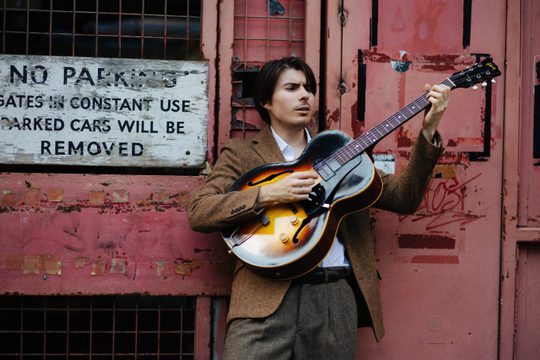 A young, white, brunette man standing in front of a red wall, playing the guitar. He is wearing a brown tweed jacket