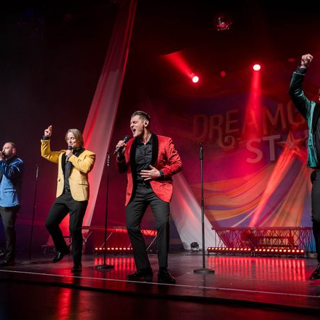 Four men in blue, yellow, red and green jackets sing on a stage. The lighting is red.