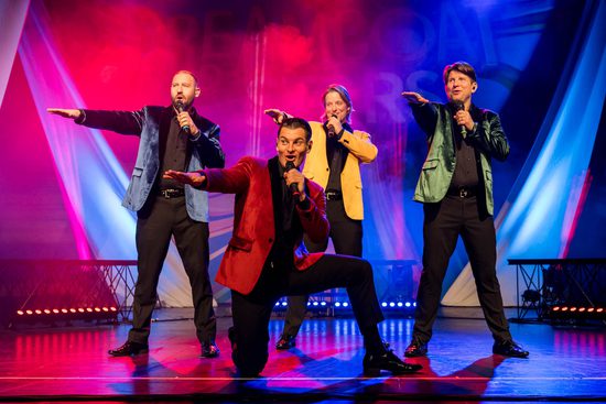 Four men in blue, red, yellow, and green jackets sing. One is kneeling on the floor and all of them have one arm straight out in front of them.