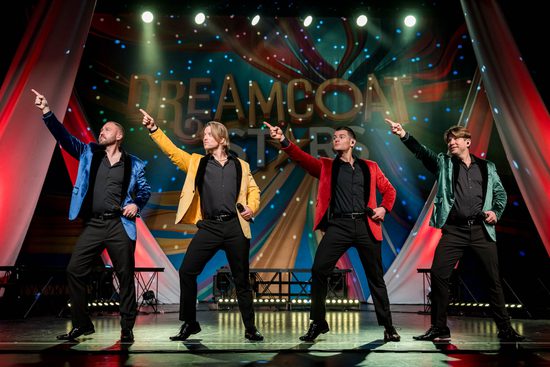 For men in blue, yellow, red and green jackets stand in a line on a stge. They are all dancing with one hand on their hip and the other pointing up. There are disco lights around them.