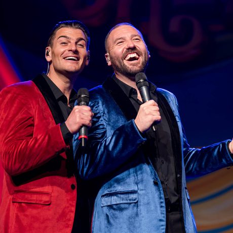 Two men, one in a red jacket and one in a blue jacket, smile with each other and hold microphones.