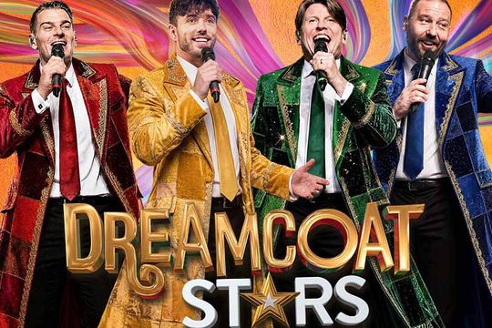 Four men in brightly coloured coat of red, yellow, green, and blue, sing into microphones in front of a multicoloured background.