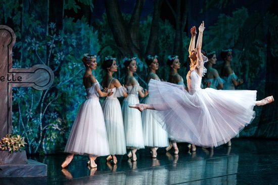 A group of women in white dresses stand in a line. In front of them, one woman in a matching dress leaps across the stage.