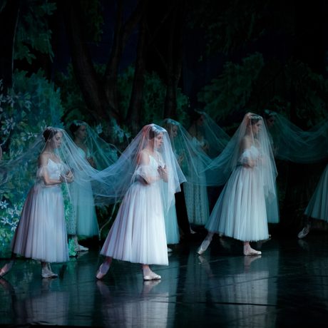 A group of women in white dresses stand on a stage next to a large cross that says "Giselle!. They are wearing white veils over their heads.