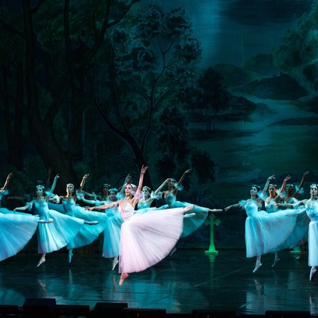 A group of women in white dresses jump with one leg out to the side. They are lit with blue light apart from one woman.