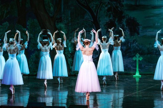 a large group of women in white dresses with flowers in their hair dance en pointe with their arms above their heads. They are lit in a teal light.