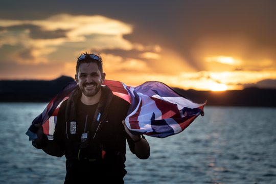 Jordan Wylie, a white man with dark hair and stubble smiles at camera with a union jack flag draped over his shoulders, behind him is the sun setting over the ocean.