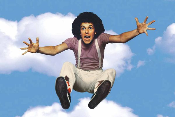 A younger Leo Sayer in a pink t-shirt and white suspenders jumps in the air. The background is a blue sky with white clouds.