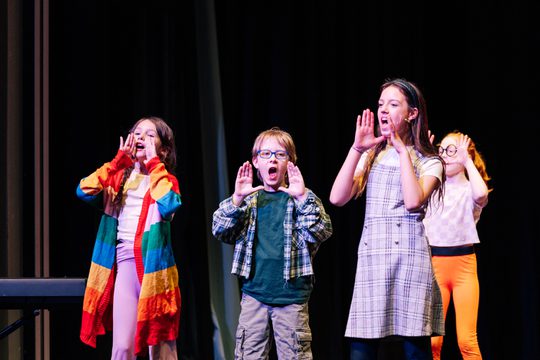 A group of children singing with their hands either side of their mouths. They are all wearing colourful outfits.