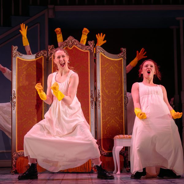 Two women in pale night dresses celebrate while wearing yellow rubber gloves. Other rubber gloved hands are poking above a burnt orange privacy screen