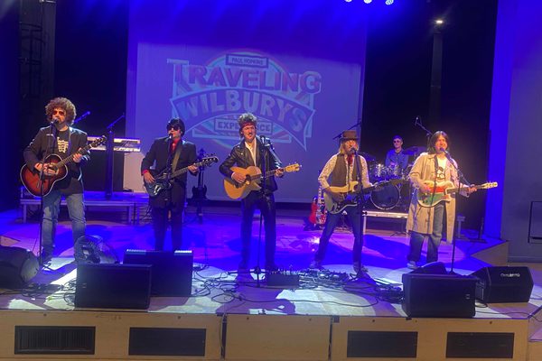 Five men on a stage. They are standing behind microphone stands and all are playing guitars.