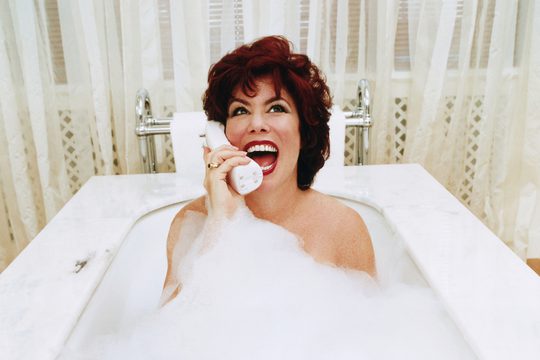 A white woman with short, red hair and glamorous make up sits on the phone in a bathtub full of bubbles.