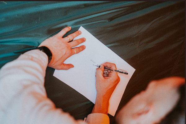 A man writing on a piece of white paper. The table is dark green.