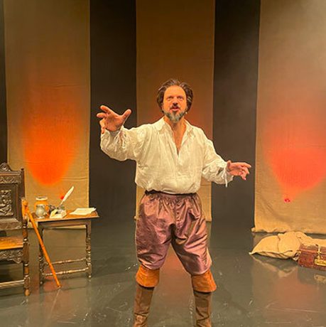 A brunette man stands on a stage next to a wooden chair. He wears a loose white shirt, purple trousers and brown knee-high boots.