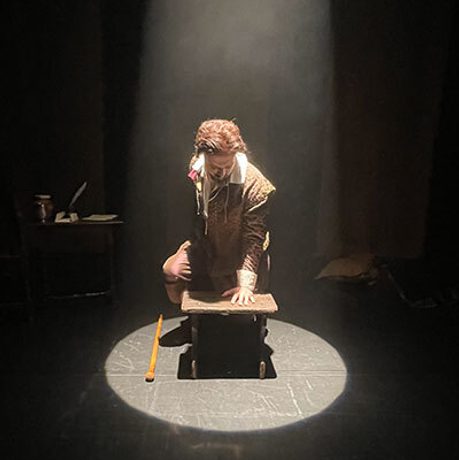 A brunette man kneels in a spotlight, with one hand on a small stool.