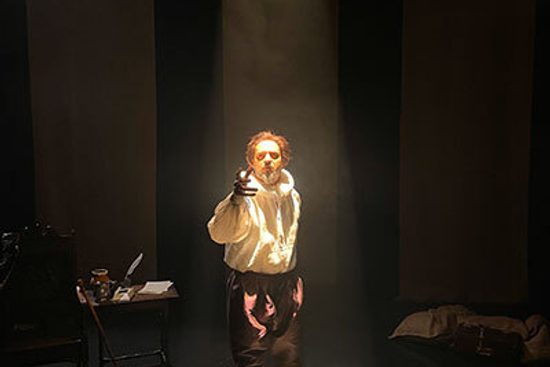 A man in a loose white shirt and neck ruff stands in a spotlight and reaches out with one hand.