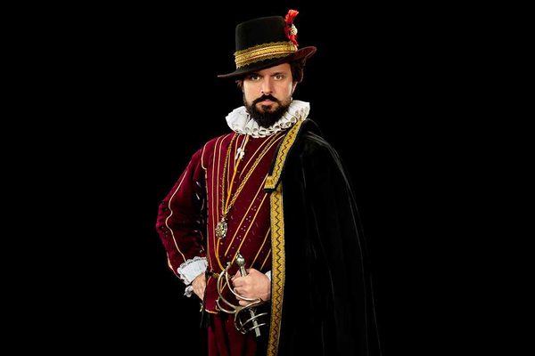 A man in an elaborate red and gold jacket, with a black cape and hat, holding a sword. He looks serious and stands against a black backdrop.