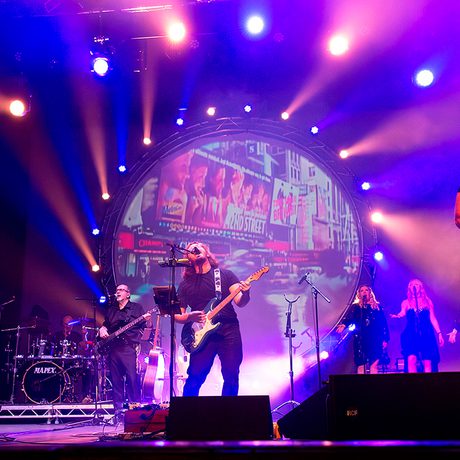A band wearing black on stage. They are playing various instruments including guitar and saxophone, and the lights and pink and blue.