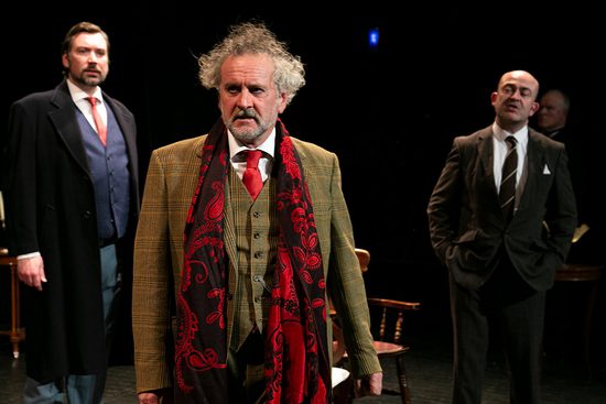 A man in a green tweed suit with a black and red scarf stands in front of two men in darker coloured suits.