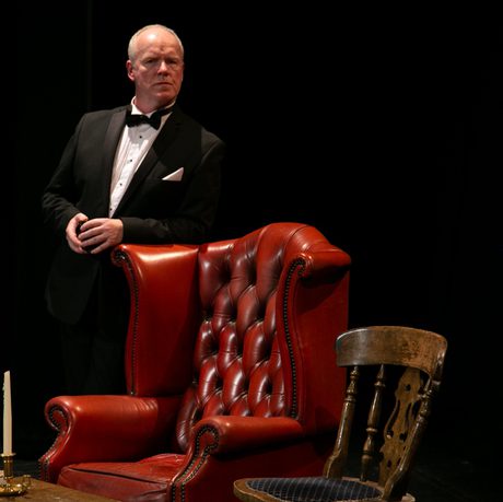 A man in a black suit and white shirt leans against a red leather chair.