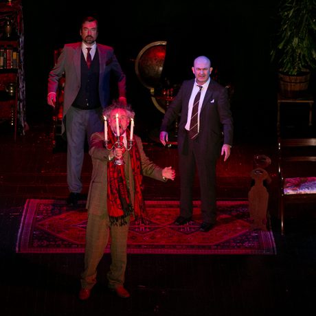 Four men in suits and one woman in a black dress stand in a living room and look up with scared expressions. One man at the front is holding a candle up to the sky.