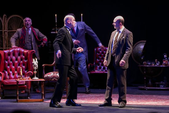 Four men in suits look at though they are arguing. They are in a living room with red leather chairs and a bar cart.