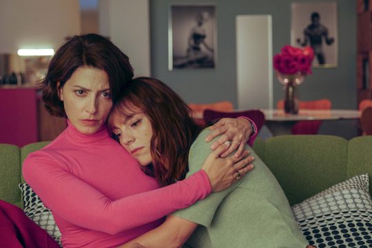 A woman in a bright pink shirt embraces a woman in a green shirt. They both look sad, and are sat on a green sofa.