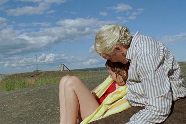 An older woman embraces a young girl. They are sat on the floor by a body of water.