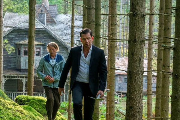 Two men walk away from a cabin into the woods. The man at the front wears a black suit, and the man at the back wears a blue raincoat.