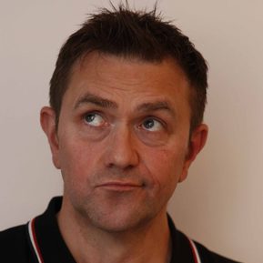 A white, brunette man wearing a black shirt looks upwards, as if deep in thought.