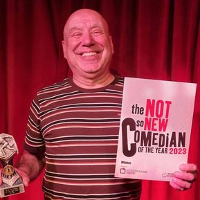 A bald, white man wearing a stripy t-shirt stands in front of a red curtain, holding an award and smiling.