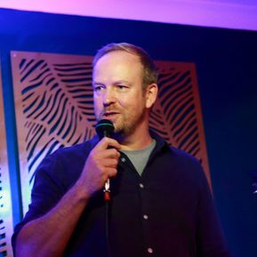 A white man wearing a navy blue shirt speaks into a microphone.