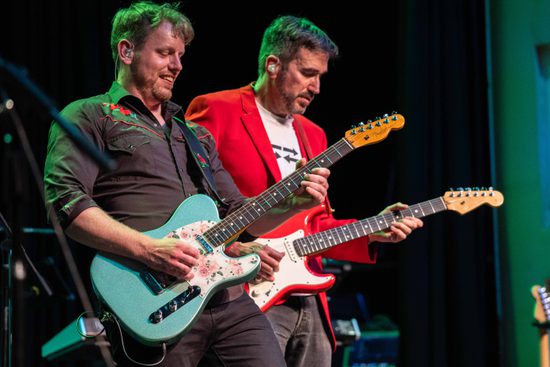Two men playing the guitar. One is in a black shirt playing a green guitar, and the other is in a red jacket playing a red guitar.