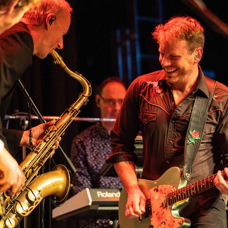 A man playing the guitar is smiling at a man playing the saxophone.