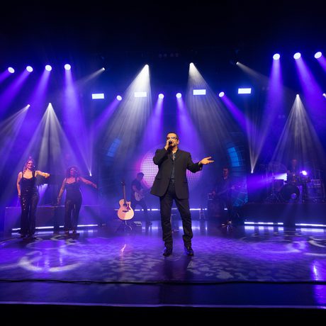 A man in a black suit on a stage with various purple spotlights around him.