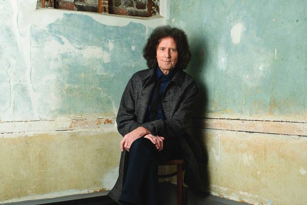 A man with brown curly hair sits against an old wall. The painted colours of yellow and blue are fading.