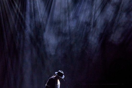 A man wearing a wide-brimmed hat on a stage. He has multiple spotlights on him.